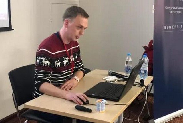 Иван Голунов сидит в свитере с оленями за столом,на котором стоит ноутбук и вода