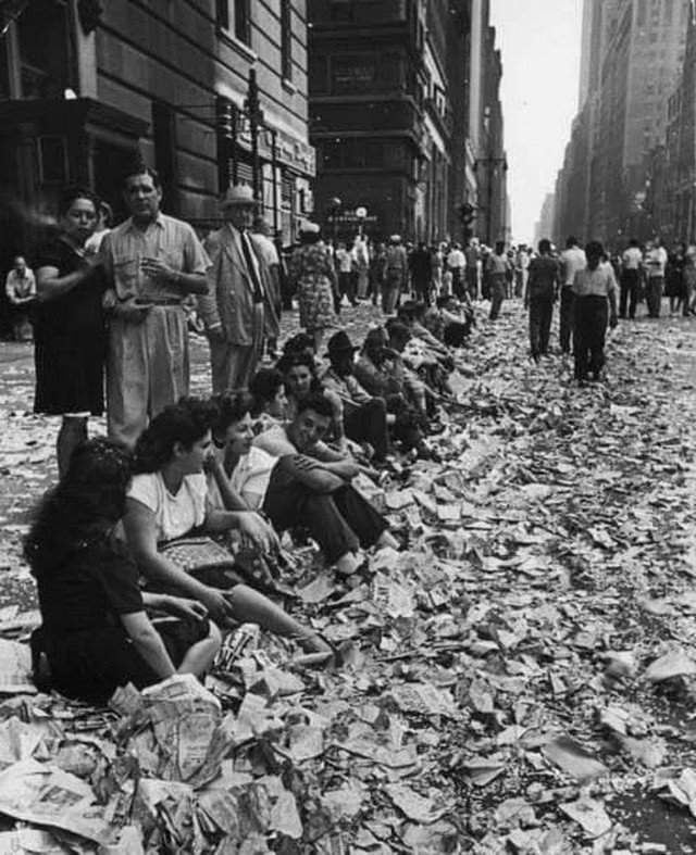 Люди, сидящие на бордюре среди конфетти и бумаг после празднования окончания Второй мировой войны в Нью - Йорке 14 августа 1945 года