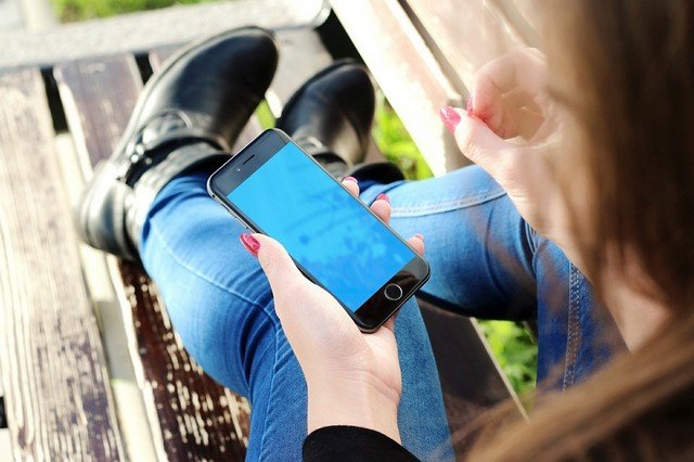Девушка держит смартфон в левой руке. Она одета в джинсы и синие ботнки, а на руках у нее красный маникюр