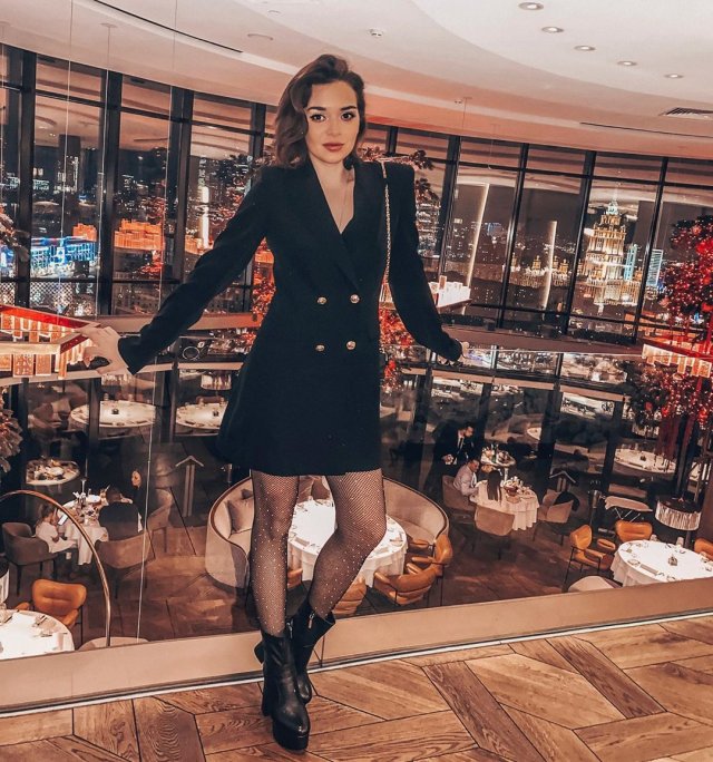 Аделина Сотникова в черном пиджаке, на фоне столов в ресторане и панорамного вида на Москву