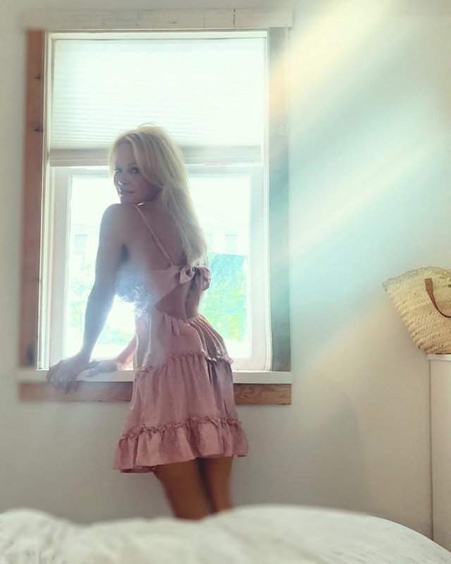 Памела Андерсон в розовом платье на фоне окна