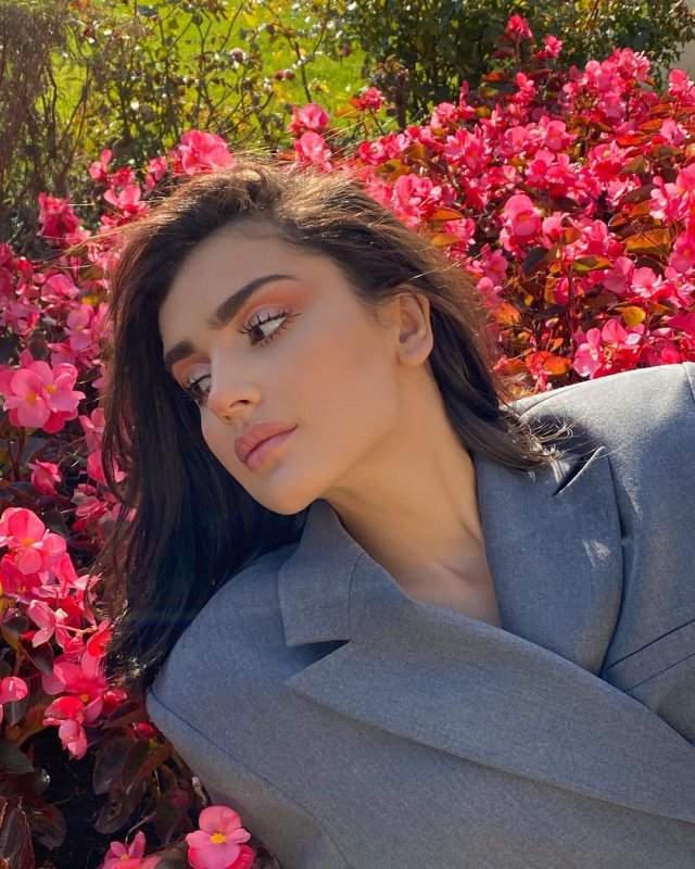 Дина Саева в сером пиджаке на фоне цветов