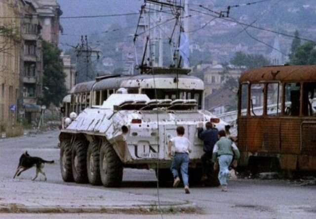 Три мальчика бегут за украинским бронетранспортером ООН, проезжающим мимо сгоревшего трамвая на площади Скендерия в осажденном Сараево 10 августа 1993 года.