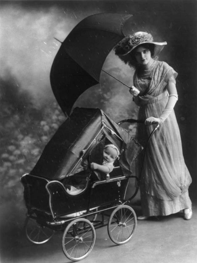 Женщина с зонтиком везёт ребенка в коляске с навесом от дождя, 1913 год